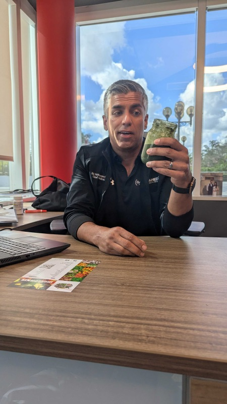 A man sits at a desk holding a rock, with papers, a laptop, and a cup nearby. Large windows and a red pillar are in the background.