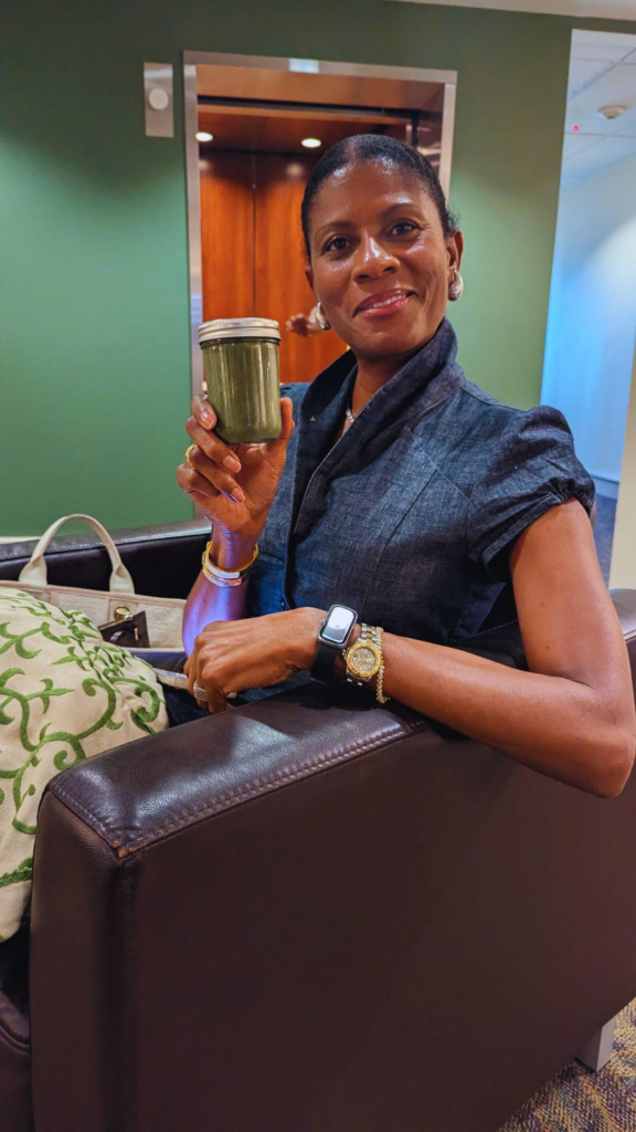 A woman sits on a brown couch holding up a jar of green smoothie, with a patterned pillow and a beige bag beside her.