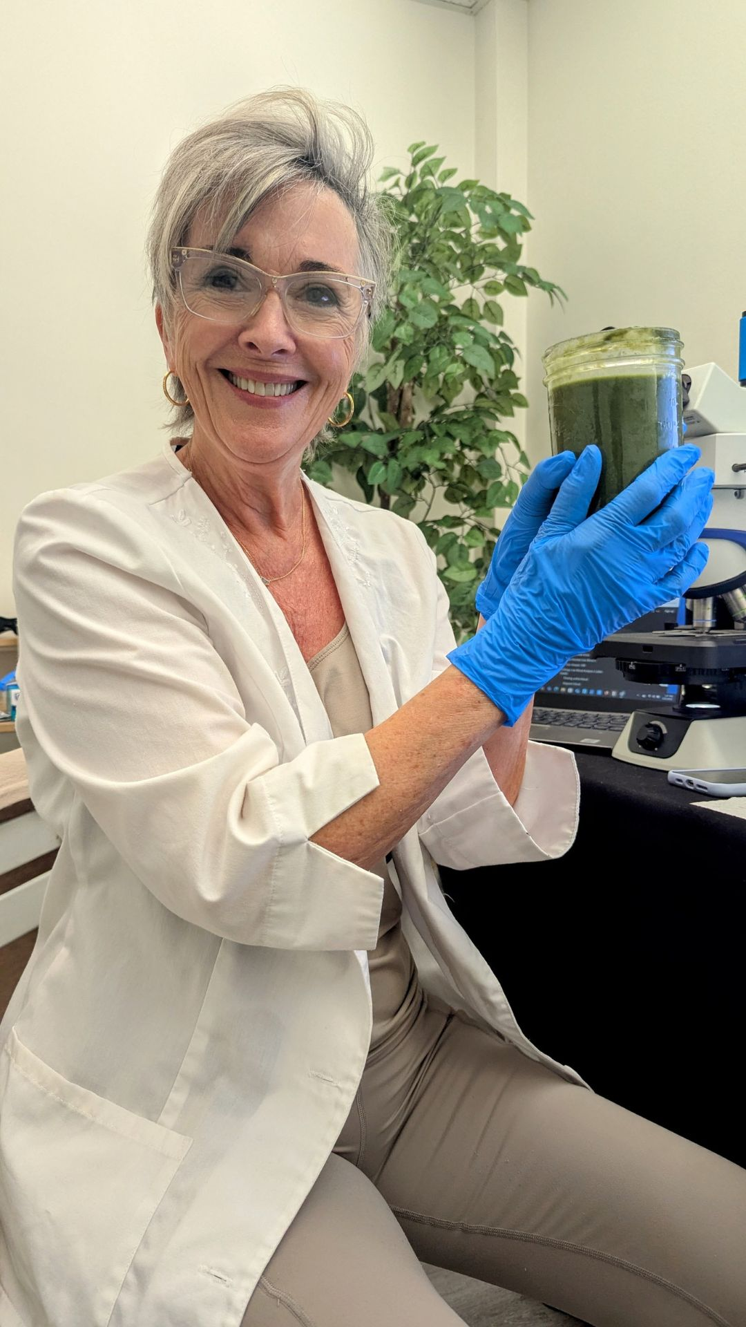 A woman wearing safety glasses and blue gloves holds a jar of green substance in a laboratory setting, with scientific equipment and a plant in the background.