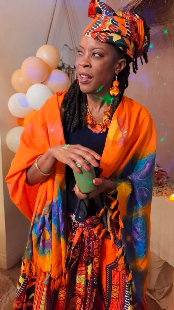 A woman dressed in vibrant, colorful traditional attire holds a green drink, standing indoors near a cluster of white and yellow balloons.