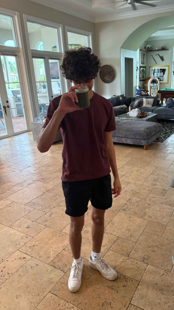 A person in a maroon shirt, black shorts, and white sneakers stands indoors, holding a glass with a green drink in front of their face.