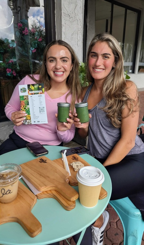 Two women sitting at an outdoor table hold green smoothies and a menu, with coffee cups and empty wooden trays in front of them.