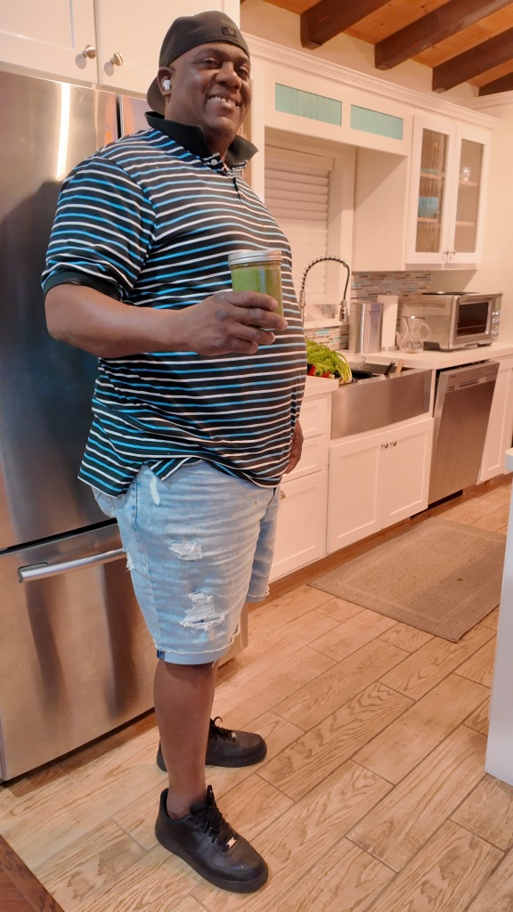 Man standing in a kitchen, smiling and holding a glass of green beverage. He is wearing a striped polo shirt, denim shorts, black sneakers, and a backwards cap.