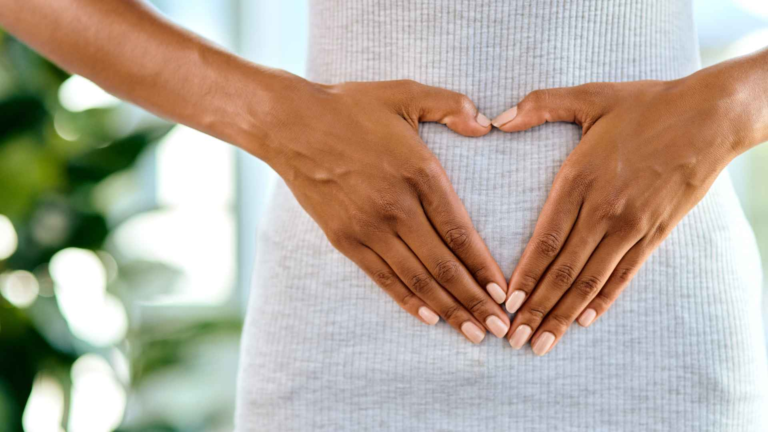 Person forming a heart shape with their hands over their stomach, wearing a light gray ribbed garment.