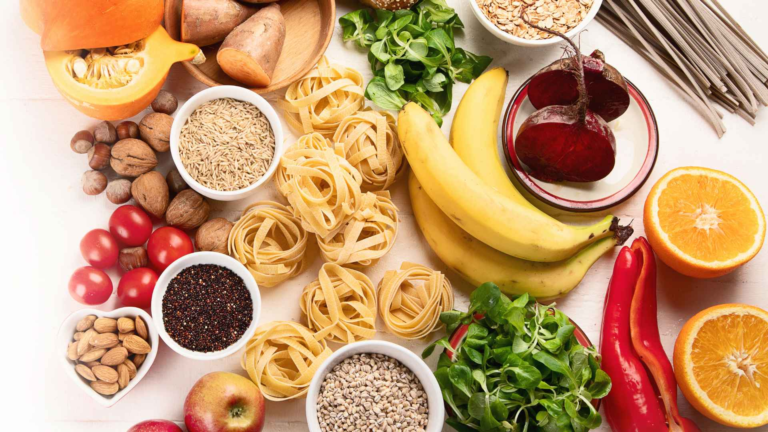Various fresh fruits, vegetables, pasta, grains, and nuts are arranged on a white surface. Foods include bananas, tomatoes, oranges, pasta nests, beets, and leafy greens—all thoughtfully selected as part of The Plate Pairing Guide for balanced meal inspiration.