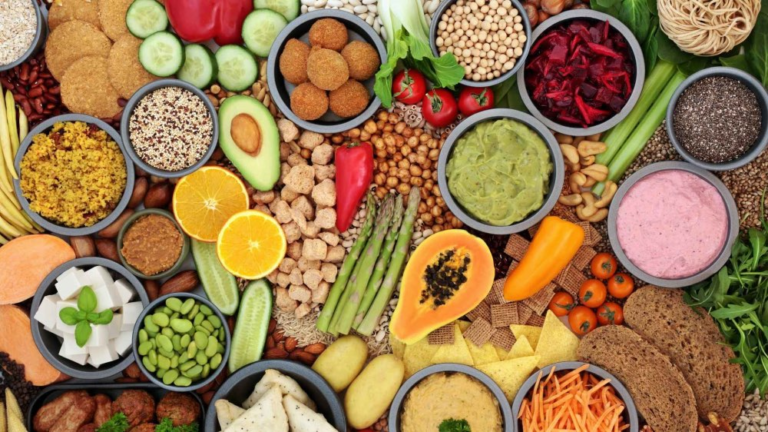 Assorted plant-based foods, including fresh vegetables, fruits, dips, legumes, grains, bread, and seeds—perfect for anyone aiming to enjoy 30 plants a week—arranged beautifully in bowls and on a table.