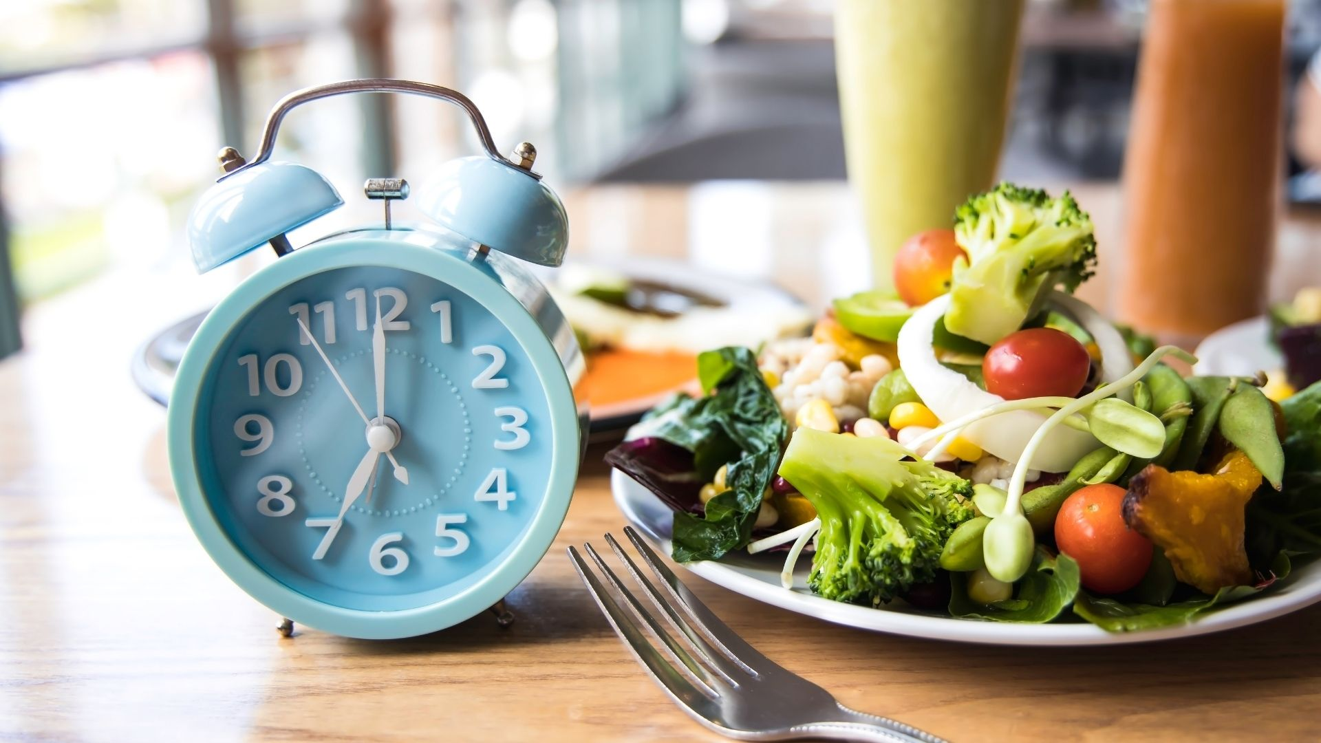 A blue alarm clock next to a plate of fresh salad with vegetables sits on a wooden table with a fork in front.