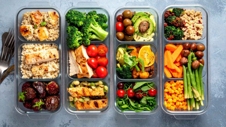 Five meal prep containers filled with assorted foods, including chicken, meatballs, broccoli, tomatoes, grains, avocado, salad greens, carrots, green beans, olives, sweet corn, and oranges.
